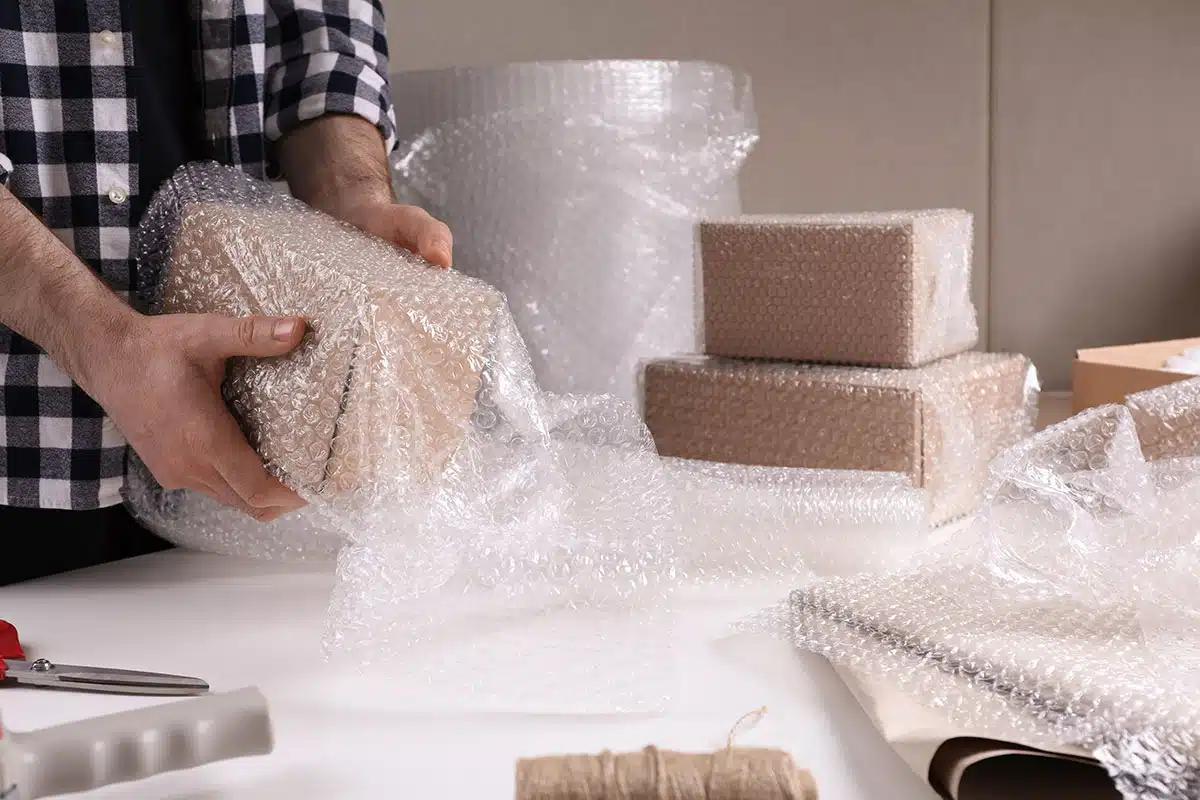 Worker hand-wrapping a fragile carton at a packing station surrounded by bubble wrap rolls and previously wrapped boxes
